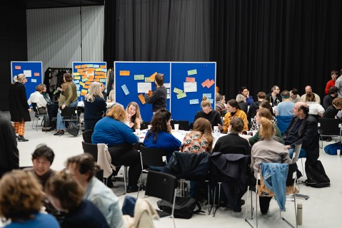 A group of people is sitting at tables working on group projects; some are standing by bulletin boards with Post-ist