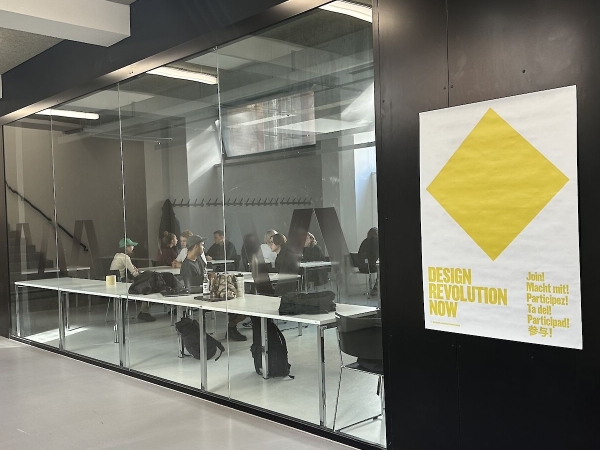 Blick durch eine Glaswand auf einen Workshopraum mit mehreren an Tischen sitzenden Personen; rechts hängt
                                          ein Plakat mit gelbem Rautensymbol und dem Schriftzug „DESIGN REVOLUTION NOW“.