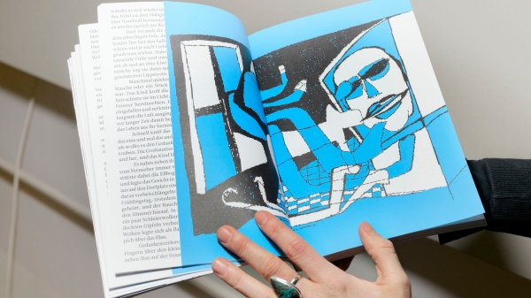 A person is holding an open book in their hands. The right-hand spread features a stylised illustration in blue, black
                                          and white: a figure is sitting in front of a computer screen. The left-hand pages contain body text. The person’s hand is
                                          wearing a striking ring with a turquoise stone.