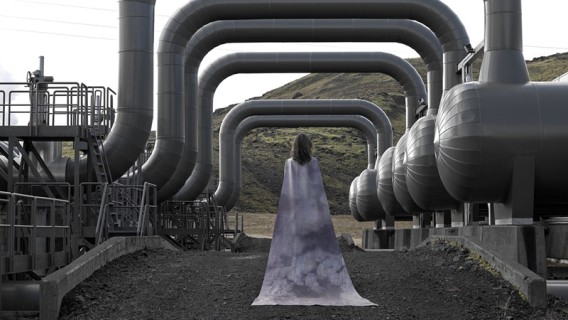 Frau mit Umhang steht in industrieller Umgebungen, die symbolisch für geologische
                           Folgen stehen