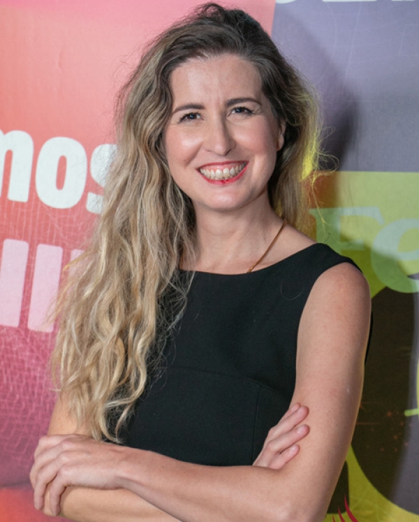Portrait of a Karla Pinhel Ribeiro smiling with long, wavy blonde hair wearing a black top. She
                                          stands with her arms crossed in front of a colorful, graphic background and looks directly at the camera.