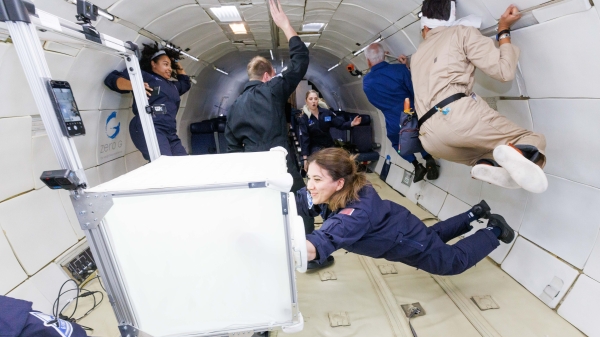 © Steve Boxall/ZeroG/MIT SEI, Ebru Kurbak, Reinventing the Spindle (2023) People in blue and
beige flight suits float inside an aircraft cabin during a zero-gravity parabolic flight. Some hold onto the walls or equipment
while others drift freely. In the foreground, one person interacts with a white, cube-shaped piece of equipment as the rest
of the group moves in the background.