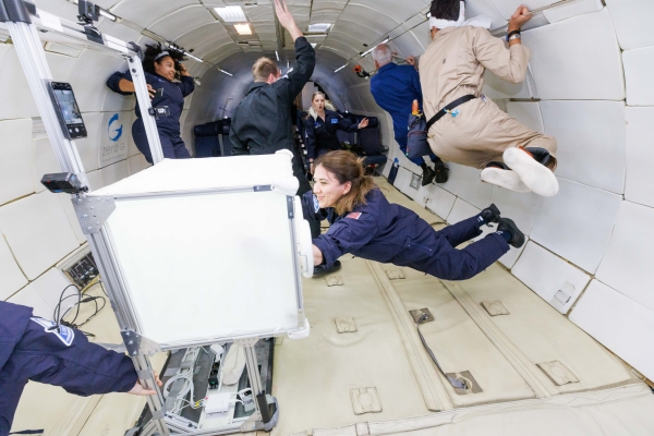 People in blue and beige flight suits float inside an aircraft cabin during a zero-gravity parabolic flight.
                                          Some hold onto the walls or equipment while others drift freely. In the foreground, one person interacts with a white, cube-shaped
                                          piece of equipment as the rest of the group moves in the background.