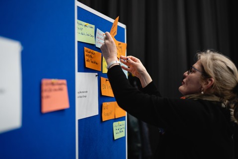 Eine Frau heftet ein Post it an eine Pinnwand bei einem Brainstorming