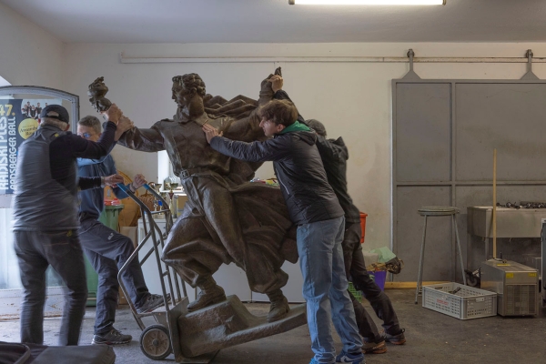 Ein Denkmal einer männlichen Figur wird von einer Gruppe Männer mit Sackkarre
                                          transportiert.