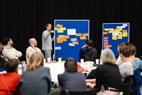 Eine Gruppe Menschen sitzt zu Gruppenarbeiten an Tischen zusammen, ein Mann steht an einer blauen Pinnwand mit Post its